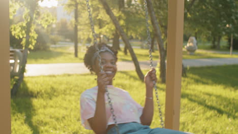 happy african american woman swinging on swing in park and posing for camera