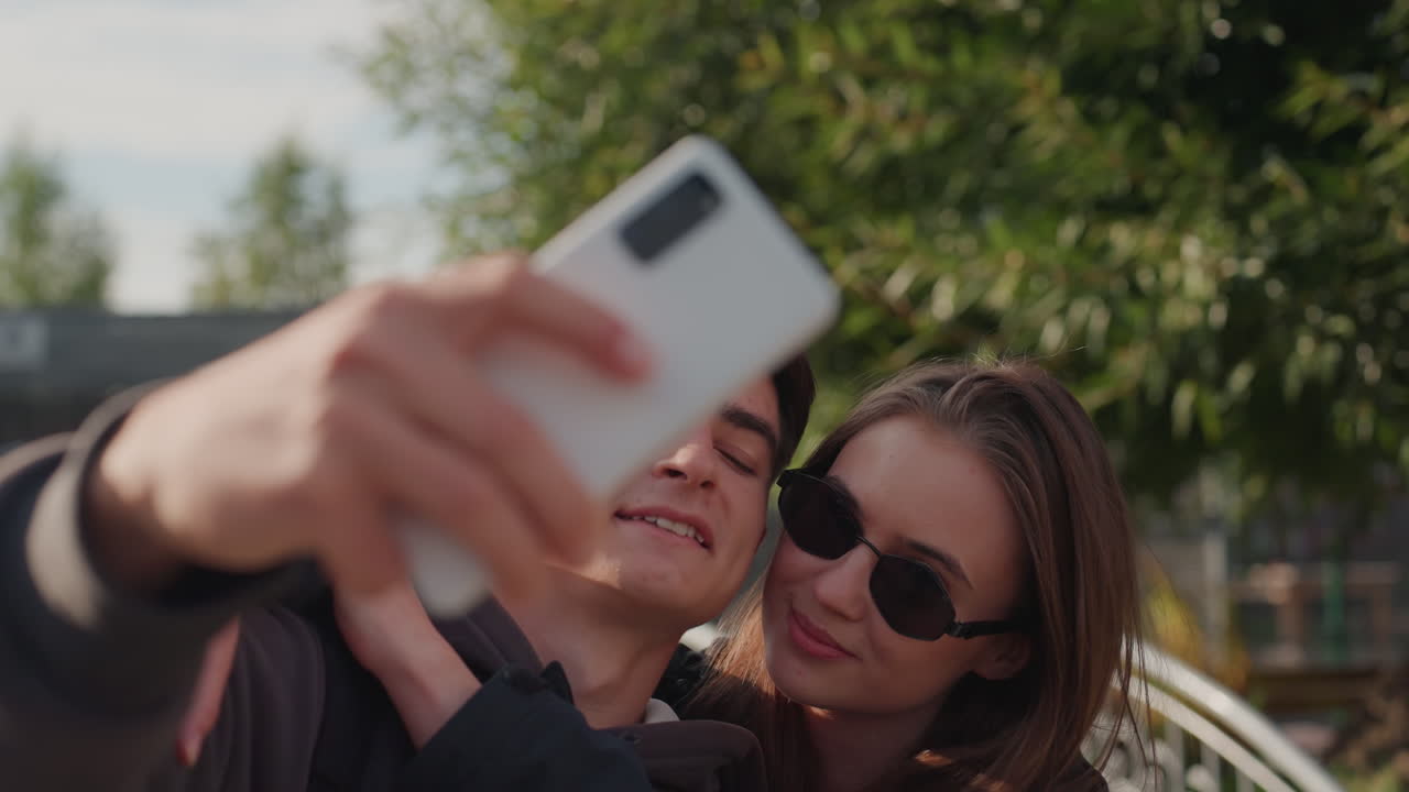 casal de adolescentes brancos se beijando durante uma selfie, abraço íntimo e close no celular sob a luz quente do pôr do sol em um jardim urbano, pose de influenciador, óculos de sol e moda casual, sorriso espontâneo, luz de fundo suave