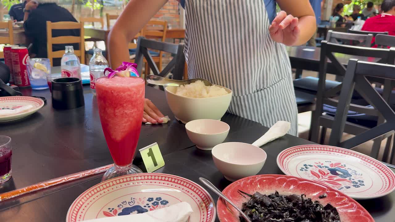 A person in striped attire serves rice at a restaurant table set with Thai seafood dishes, vibrant drinks, and decorative plates under bright indoor lighting