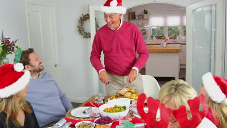 Abuelo-Tallando-Pavo-Mientras-Una-Familia-Multigeneracional-Se-Sienta-A-Comer-Juntos-La-Comida-Navideña