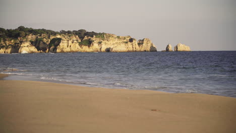 Las-Olas-Rompen-En-La-Playa-Con-Los-Acantilados-De-Alvor,-Portugal-En-El-Fondo.