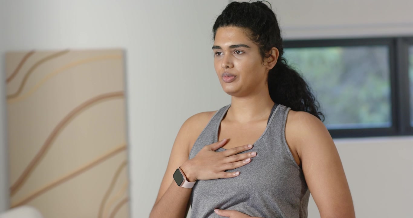 Donna indiana con le mani sul petto e sull'addome che pratica esercizi di respirazione a casa con uno smartwatch