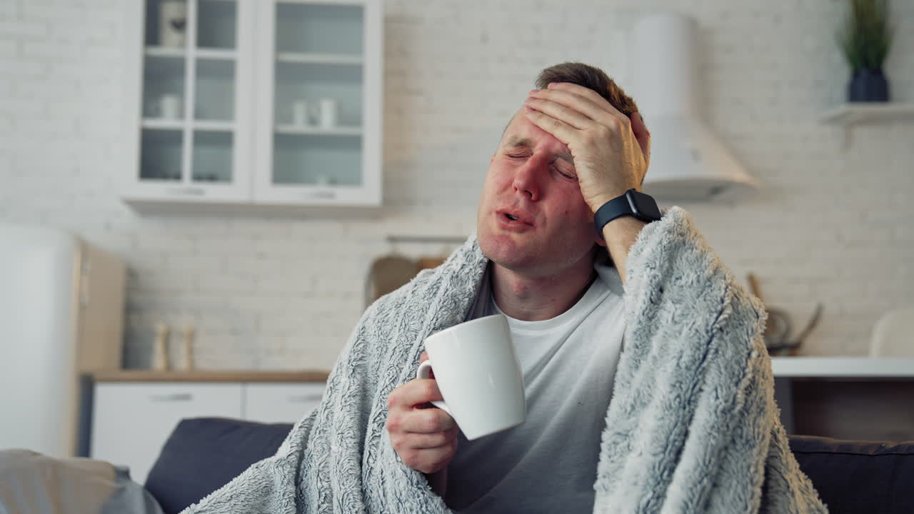 Folk medicine. Sick young man with fever wrapped in warm blanket drinking hot tea. Pandemic.