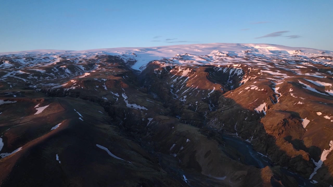 Luftfoto af landskab med bjerge og gletsjerdale, hvor sne smelter ned i floder, på Island