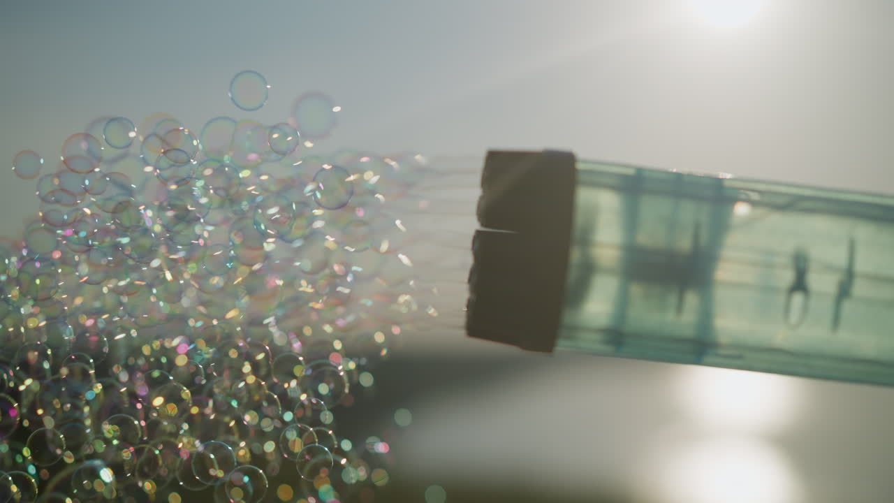 플라스틱 거품 총이 밝고 약간 구름이 많은 하늘 아래 다채로운 거품의 흐름을 방출하는 클로즈업.