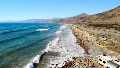 toma aérea de un dron elevándose sobre un campamento de vehículos recreativos en la playa con olas azules del océano rompiendo contra la línea costera dorada de california