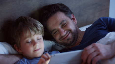 top view of happy caucasian father lying on the bed with his cute little son scrolling and taping on the tablet at night