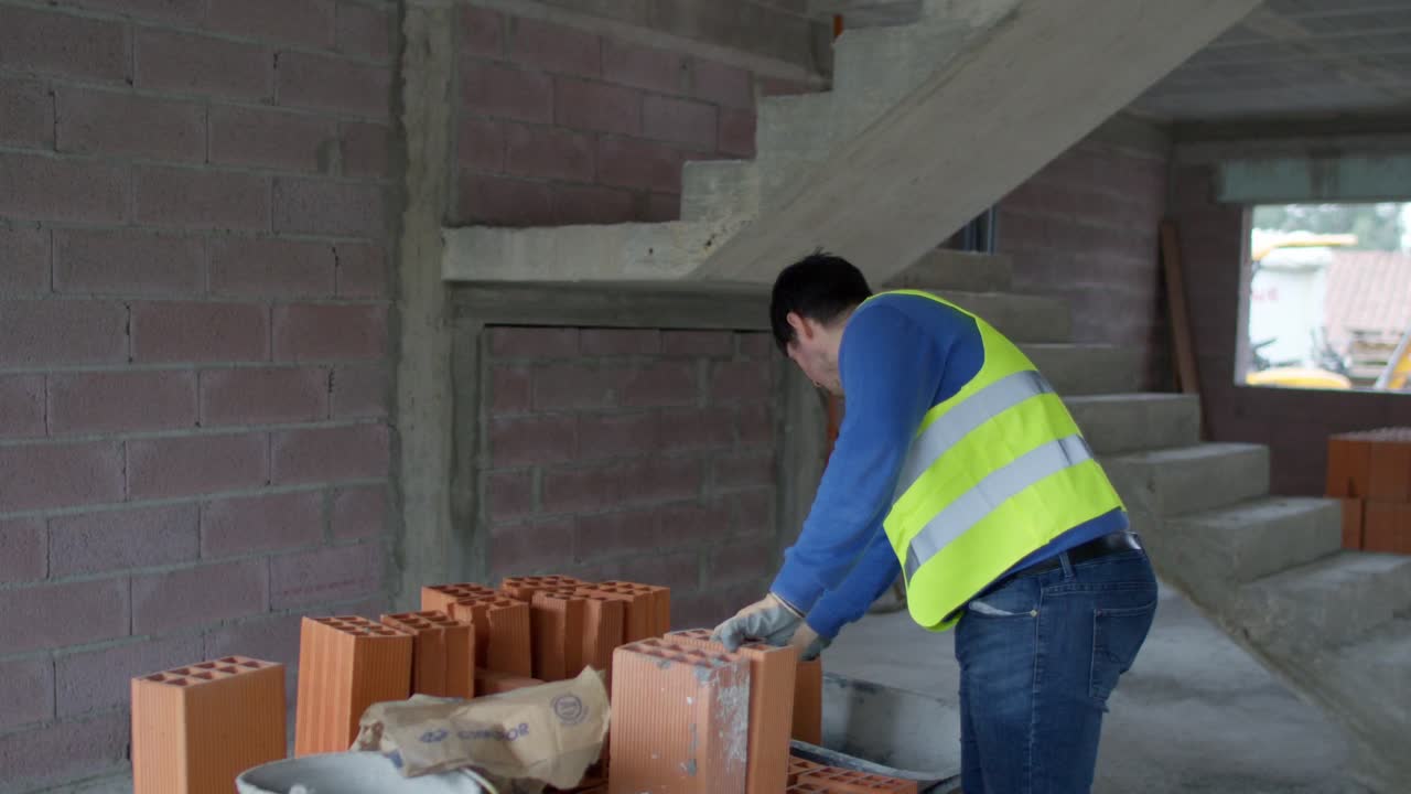 4K slow-motion footage of a worker in a safety vest loading bricks into a wheelbarrow, captured with a gimbal move from behind, showing the stack of bricks at a construction site