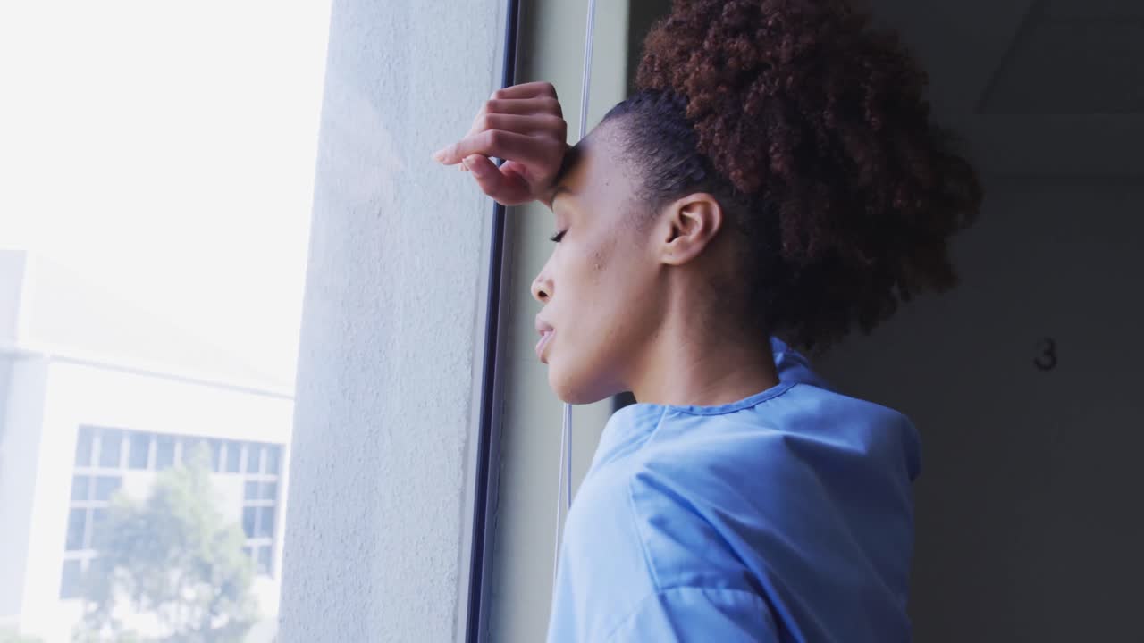 Tired mixed race female doctor standing in hospital room looking through window