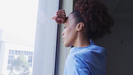 Tired-mixed-race-female-doctor-standing-in-hospital-room-looking-through-window