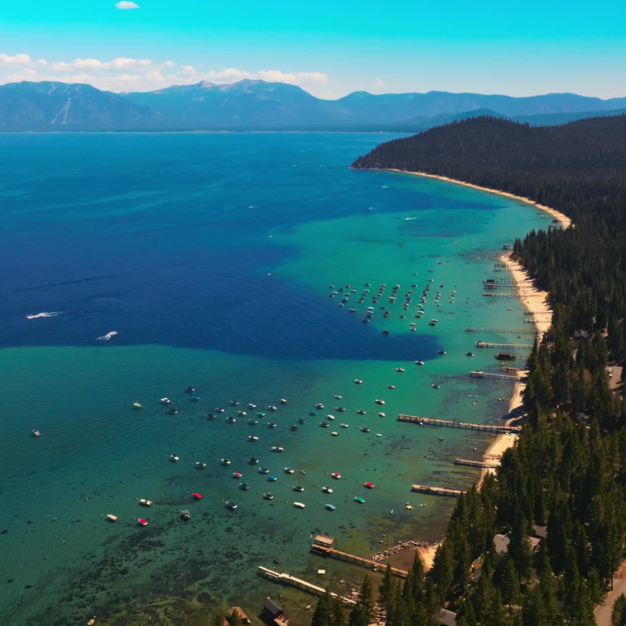 Turquoise waters of amazingly beautiful Lake Tahoe with wooded shore. The Sierra Nevada mountains at backdrop. Aerial view