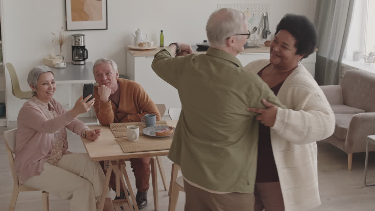 High angle of happy African woman dancing waltz with her senior Caucasian husband, their friends filming them on smartphone sitting at dinner table