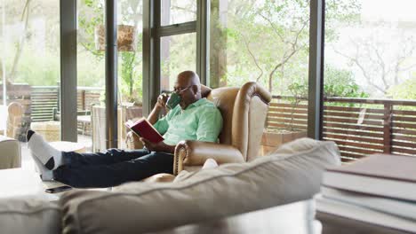 happy african american senior man relaxing in armchair, feet up, reading book and drinking coffee