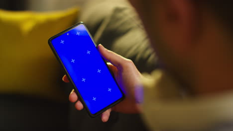 close up over the shoulder shot of man spending evening at home sitting on sofa looking at blue screen mobile phone 3