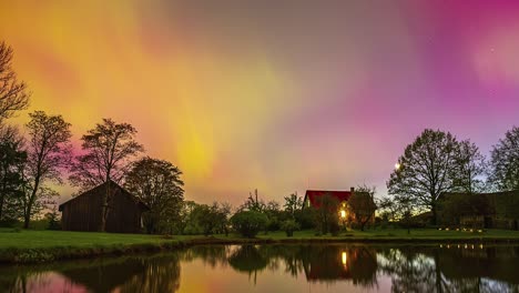 Brillante-Aurora-Boreal-Sobre-Una-Granja-Con-Los-Colores-Del-Cielo-Reflejándose-En-El-Lago---Lapso-De-Tiempo