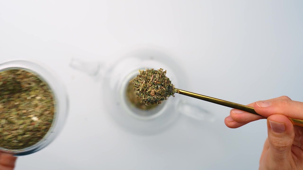 Dipping spoon in dry melissa leaves in glass jar. Top view, hands using yellow utensil inside transparent round container with green tea herbs on white backdrop.