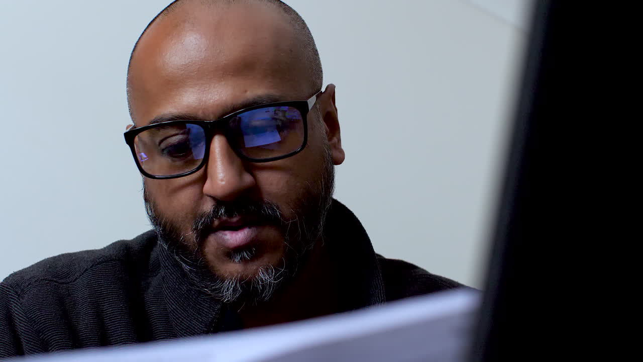 Close-up of a man with glasses, appearing anxious and despondent while reading documents, possibly related to bills or cost of living