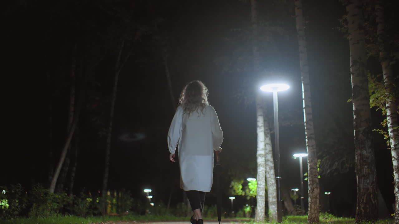 Back view of young girl in long white coat walking through peaceful park path under bright streetlights at night, holding umbrella, surrounded by trees and greenery in serene calm atmosphere
