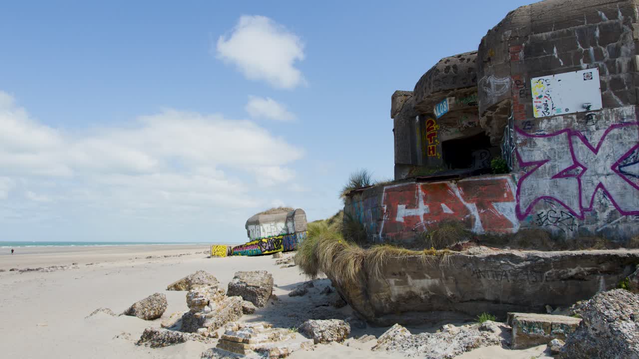 Static wide shot of graffiti-covered bunker ruins on sandy beach under bright daylight, coastline visible
