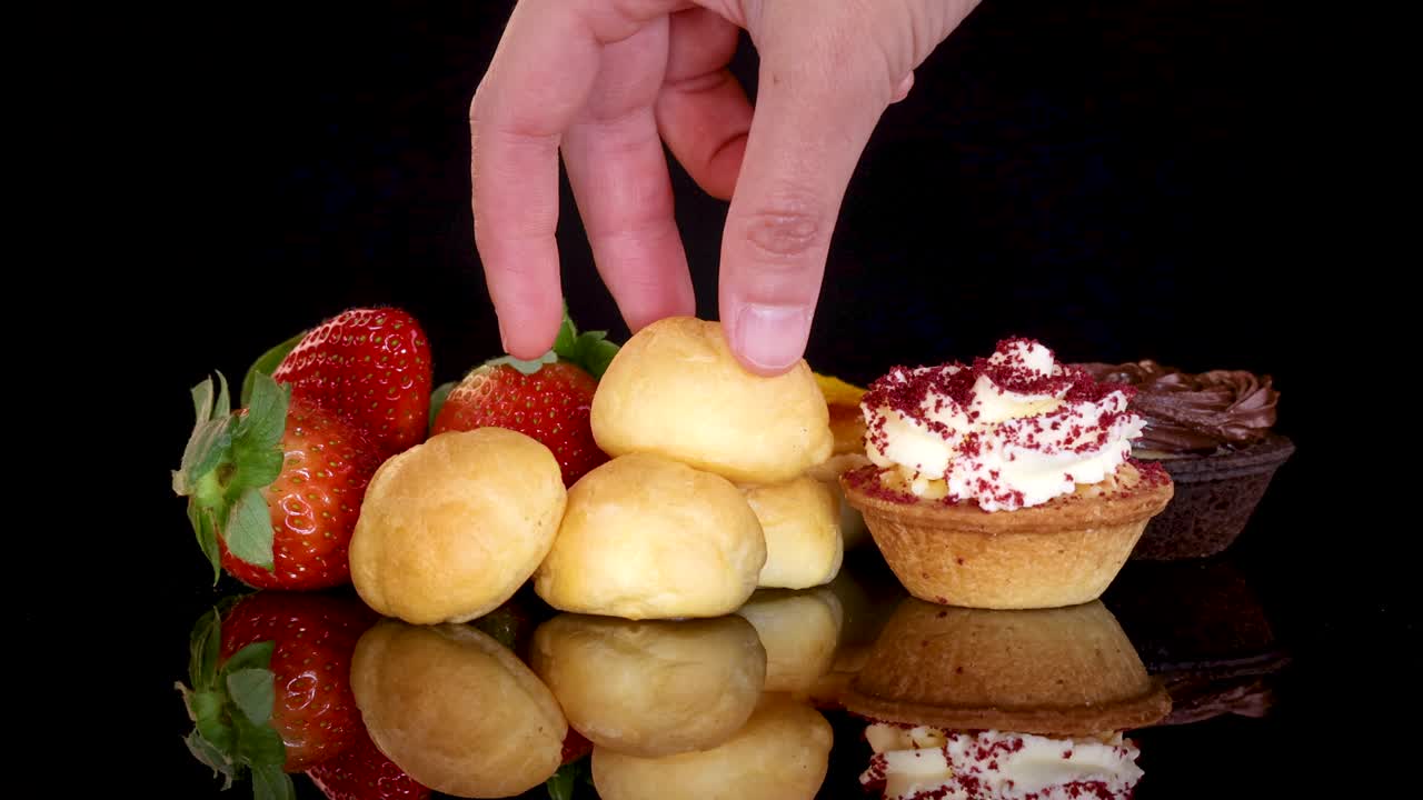 Hand selects cream puff from assorted desserts on glossy black surface with strawberries and cupcakes