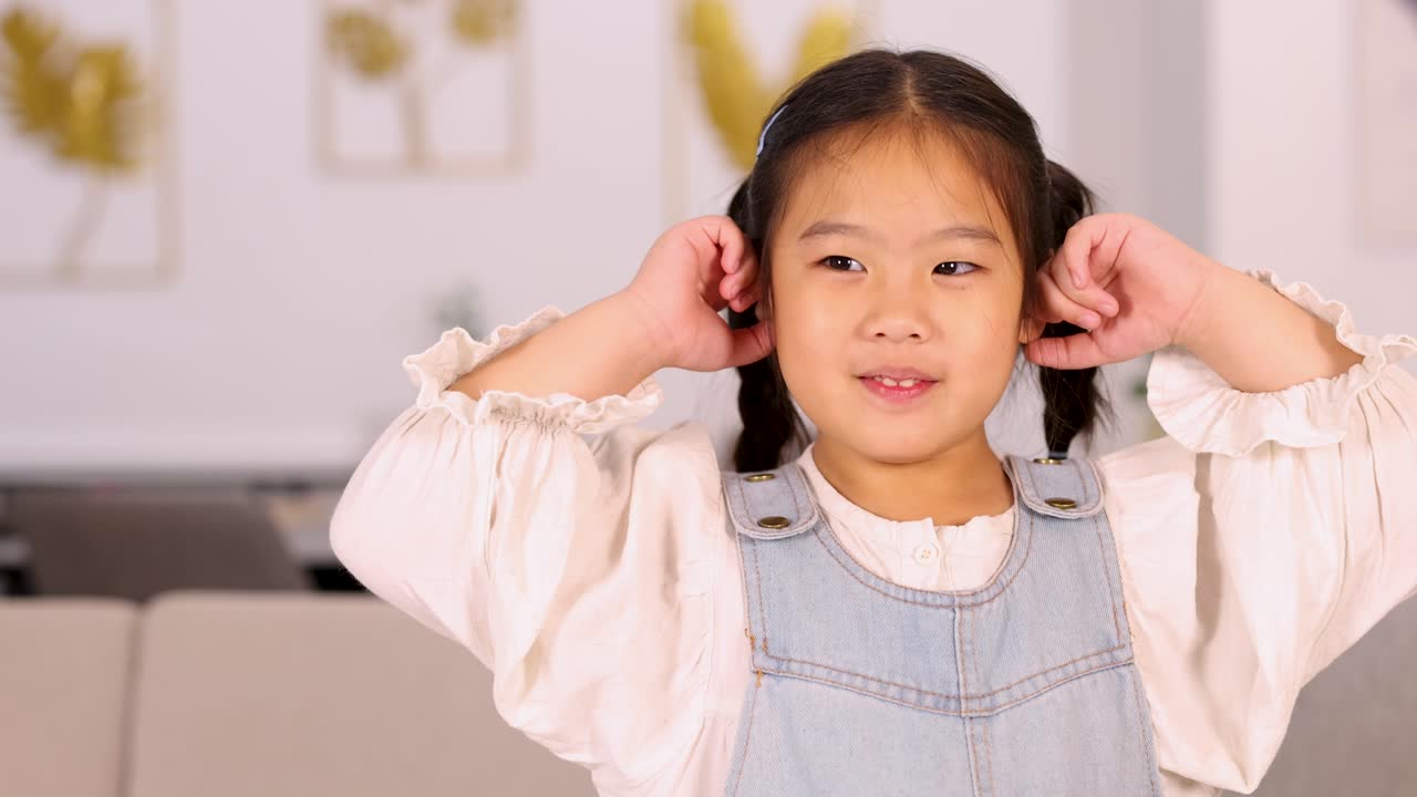 Young Asian girl covers her ears with both hands, expressing discomfort or playfulness, in a brightly lit modern living room with soft background decor