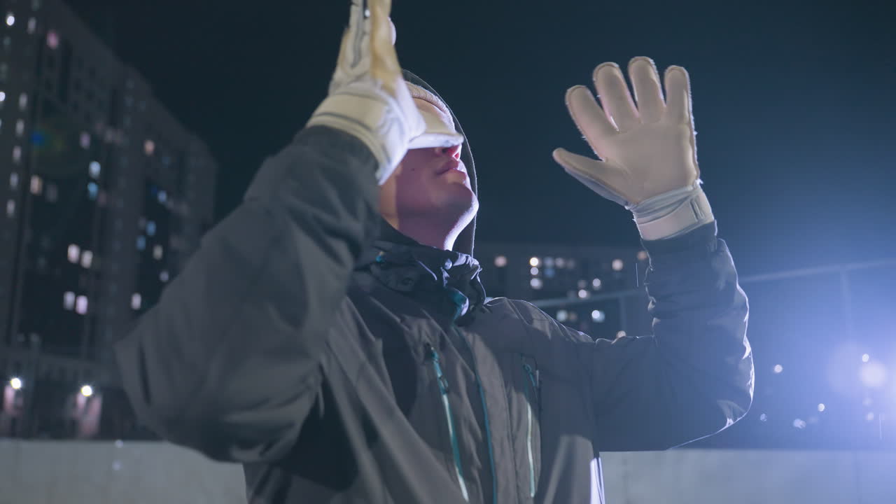 손바닥을 착용한 축구 선수가 공중으로 축구 공을 던지고 도시 필드에서 야간 훈련 기간 동안 다시 잡는 측면 뷰, 밝은 도시 불빛과 흐릿한 배경