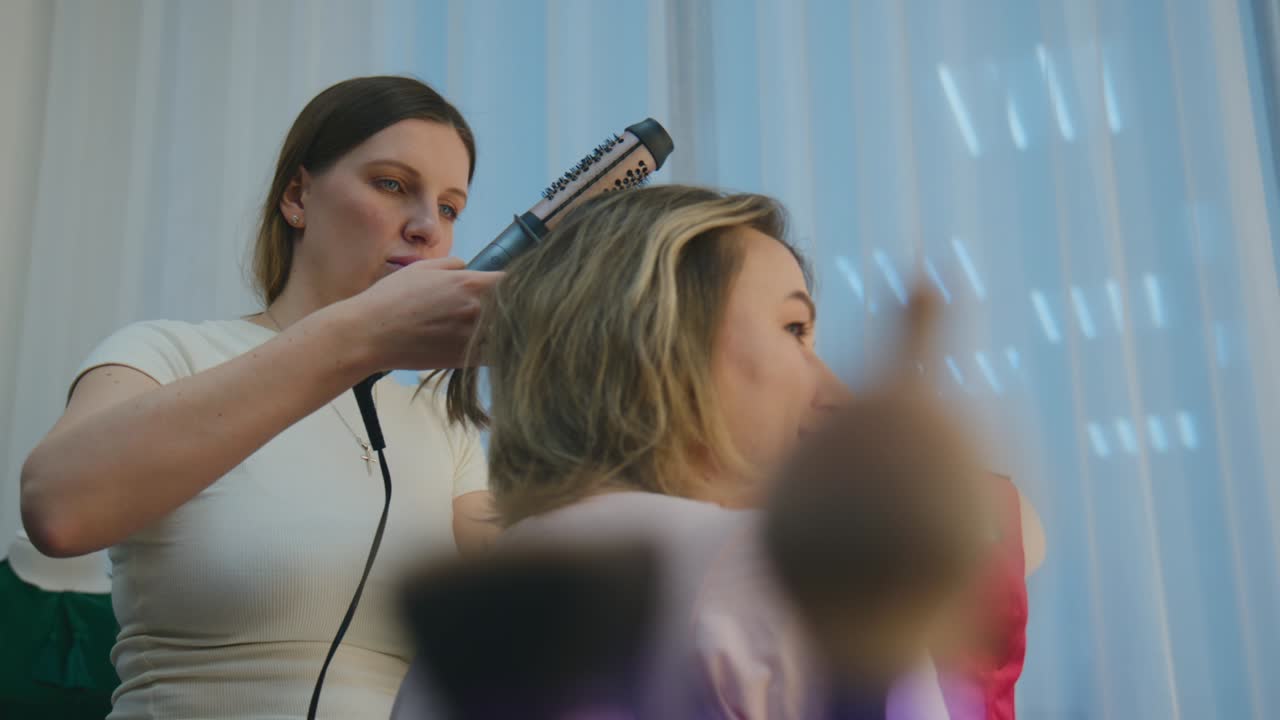 Stylist carefully uses a hairdryer on a client's hair. The stylist focuses intently on styling, showing concentration. The client sits patiently, trusting the stylist's skill