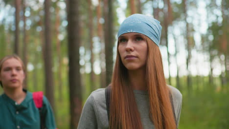 women hiking through a serene forest, woman in blue bandana and gray sweatshirt looking attentively to the side, while her companion in green shirt is softly blurred behind