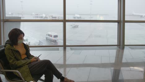 a young woman in a yellow jacket and a green raincoat and a medical mask sits alone