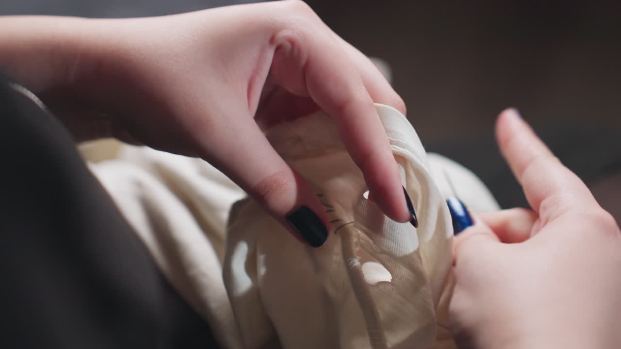 Close up hands with polished nails stitching torn white dress with needle and thread, steady work on lap, black outfit, indoor focus, careful repair of fabric seam, soft home light, precise movement