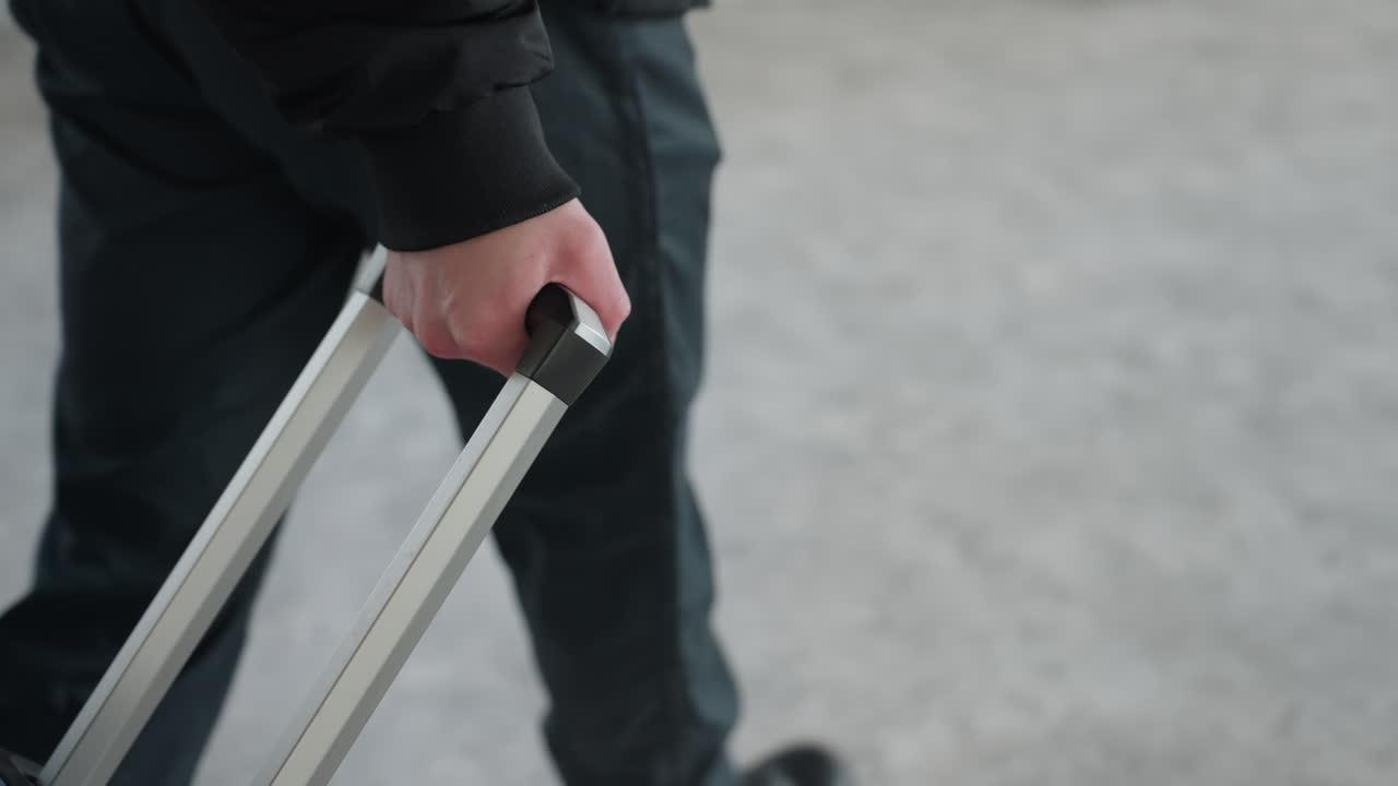 Man walking indoors pulling wheeled travel bag handle to front in slow motion on smooth concrete floor under soft daylight in neutral toned interior, casual black jacket and pant leg visible in frame