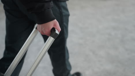 man walking indoors pulling wheeled travel bag handle to front in slow motion on smooth concrete floor under soft daylight in neutral toned interior, casual black jacket and pant leg visible in frame