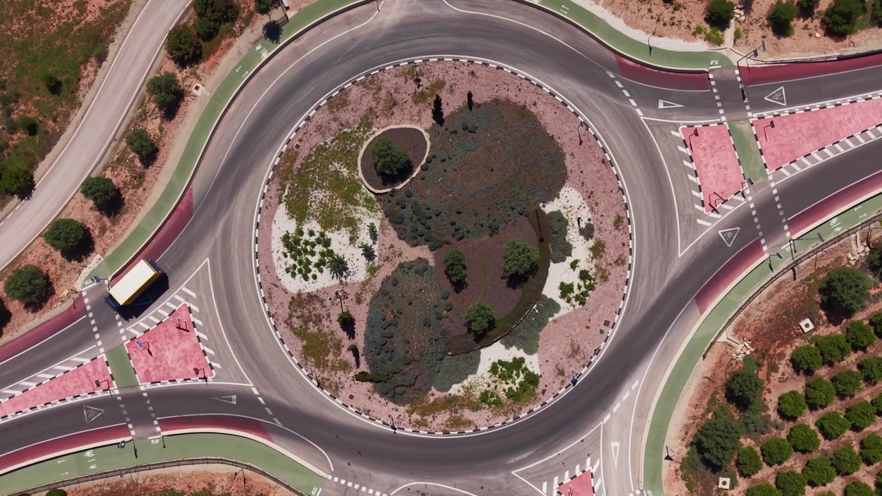 Top-down view of a large road roundabout with a landscaped central island featuring shrubs and trees, surrounded by colored pedestrian crossings and road markings