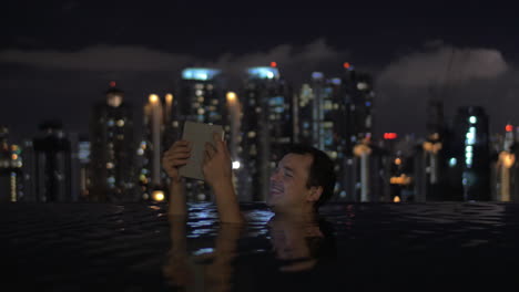 En-La-Piscina-En-El-Techo-De-Un-Hotel-En-Kuala-Lumpur,-Malasia,-Un-Hombre-Mira-Videos-En-Una-Tableta-Y-Sonríe