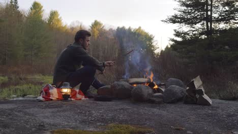 a man sits beside a campfire, cooking over an open flame at dusk