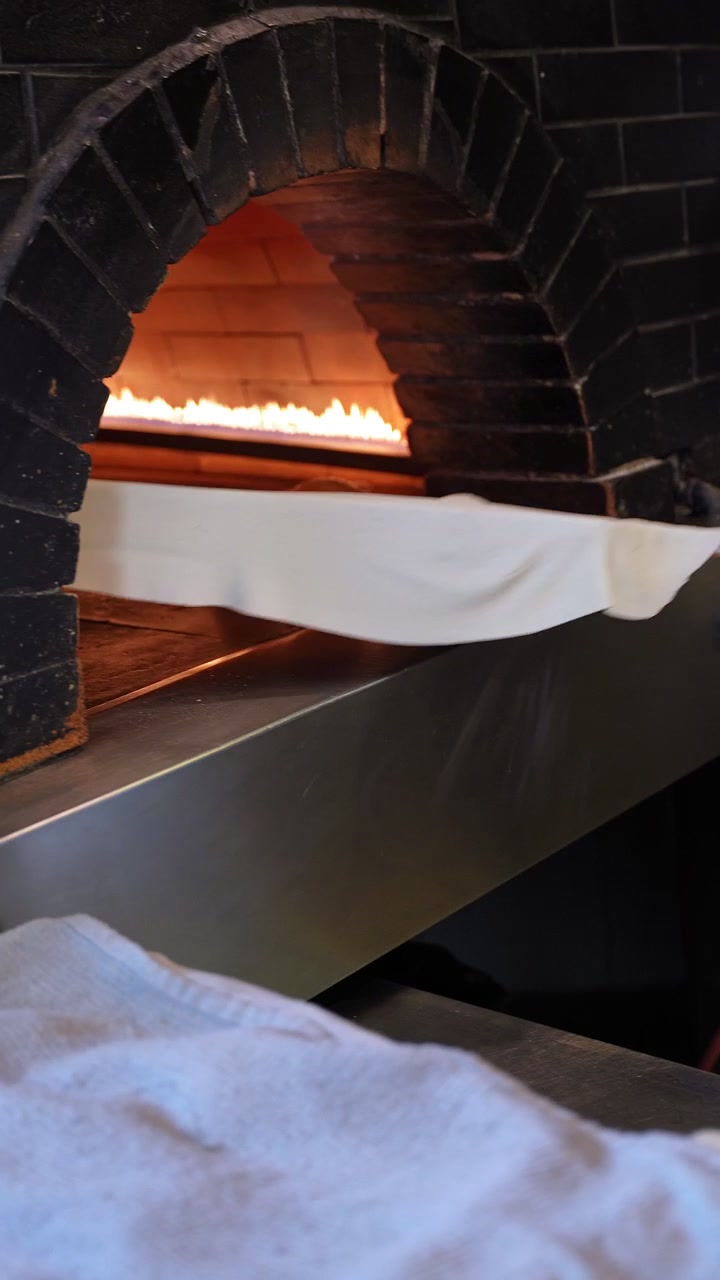 Nærbillede af rå dej på en træspade, der placeres inde i en traditionel sort stenovn med flammende ild i et restaurantkøkken, filmet i lodret smartphone-format