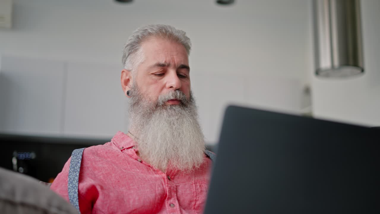 A slightly confused elderly man with gray hair and a lush beard in a pink shirt is working on his gray laptop while sitting on the sofa in a modern apartment