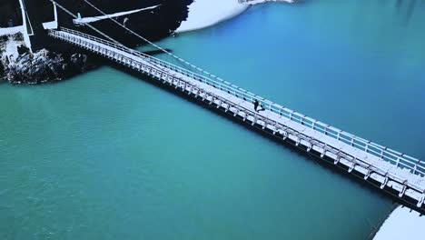 Una-Persona-Camina-Sobre-Un-Puente-Colgante-En-El-Valle-De-Skardu:-Un-Río-Con-Agua-Azul-Y-El-Dron-Se-Cierne-Sobre-El-Puente