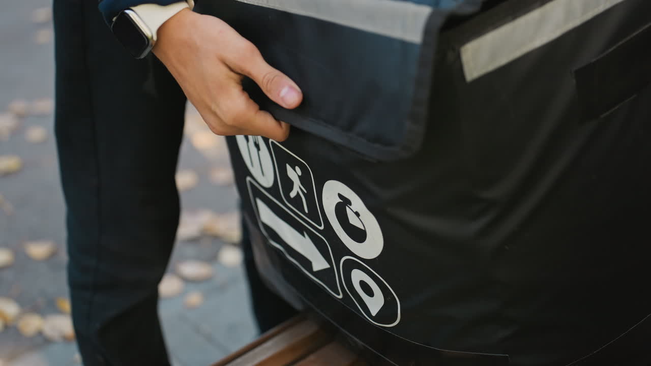 courier handling insulated delivery bag outdoors closeup, hand lifting flap to reveal food containers inside black thermal bag with white icons, reflective strip catching evening light, autumn leaves