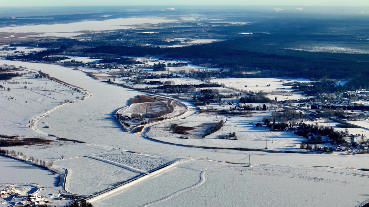 Panoramisk droneopptak fra luften fanger en fredfull vinterhistorie om en stille landsby som ligger mellom frosne elvebredder og mørke furuskoger under en klar blå himmel med skarpt kaldt vær og hvit snø.