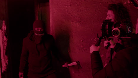 camarógrafo y trabajador de producción grabando una escena de un hombre con pasamontañas negro sosteniendo un hacha en un edificio en ruinas 1