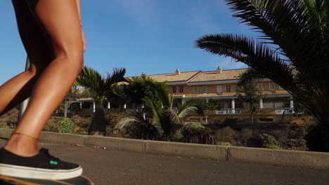 Cámara-Lenta-Enmarca-El-Viaje-En-Longboard-De-Una-Mujer-Joven-Cerca-De-Palmeras,-Su-Vestimenta-De-Pantalones-Cortos-Y-Zapatillas-De-Deporte-Refleja-El-Ambiente-Veraniego