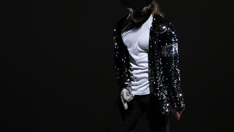 a person wearing a sparkly jacket, fedora, and sunglasses poses against a dark background