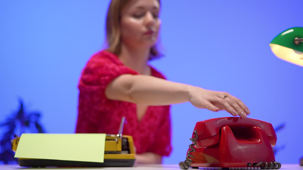 A woman in a red floral blouse speaks on a red rotary phone while seated at a desk with a yellow typewriter. The vivid blue background and retro elements create a colorful vintage-inspired setting