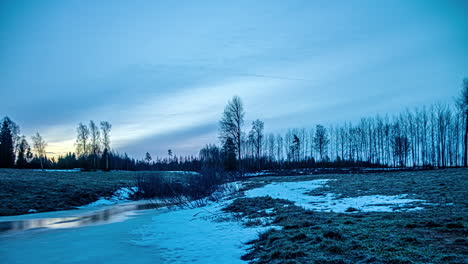 Timelapse-shot-of-frozen-snow-melting-into-a-stream-in-snowy-winter-landscape-with-sun-rising-in-the-background