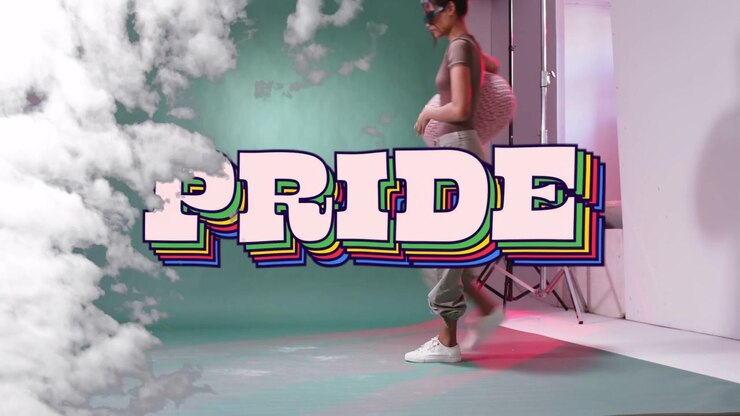 Entrando da destra del palco, un'artista donna porta un pouf, si siede e posa per celebrare il PRIDE.