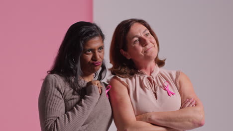 portrait en studio de deux femmes matures portant des rubans de sensibilisation au cancer du sein roses s'embrassant sur un fond rose 2