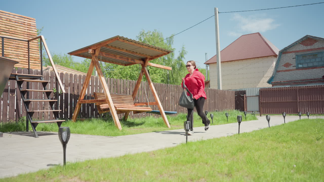 Happy lady in red jacket runs cheerfully along paved path past wooden swing under metal roof in green backyard near brown fence and suburban houses under bright sunny sky with fresh outdoor vibe