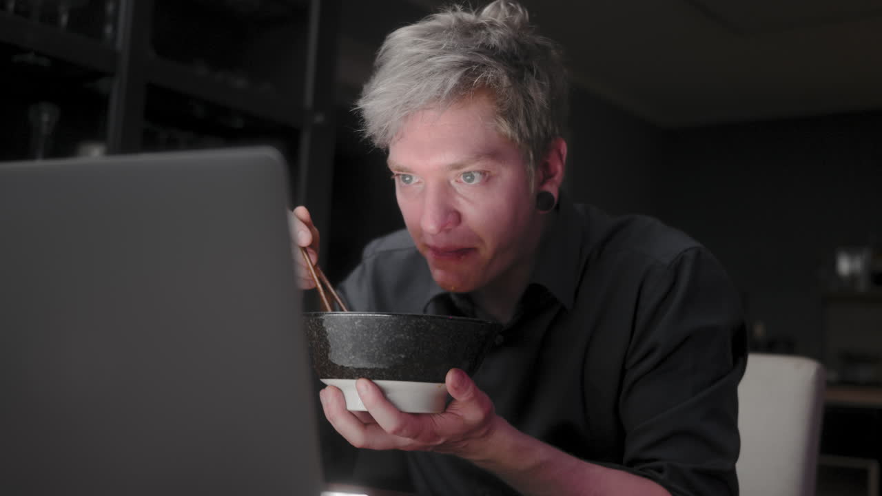 joven con el pelo plateado y un pantalones negro de negocios sentado solo en una habitación oscura comiendo sopa de fideos asiáticos y bebiendo de un tazón, mientras mira fijamente una pantalla brillante de un portátil
