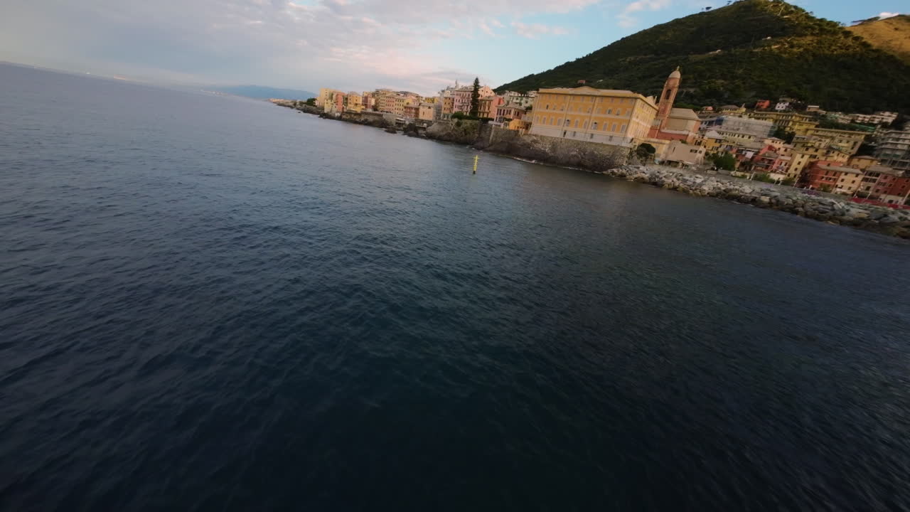FPV drone flying above Liguria coast with colorful seaside town, calm sea, buoy, mountains and golden sunset light